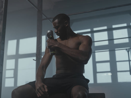 Athlete seated on a gym bench using a handheld massage gun on the thigh after a workout